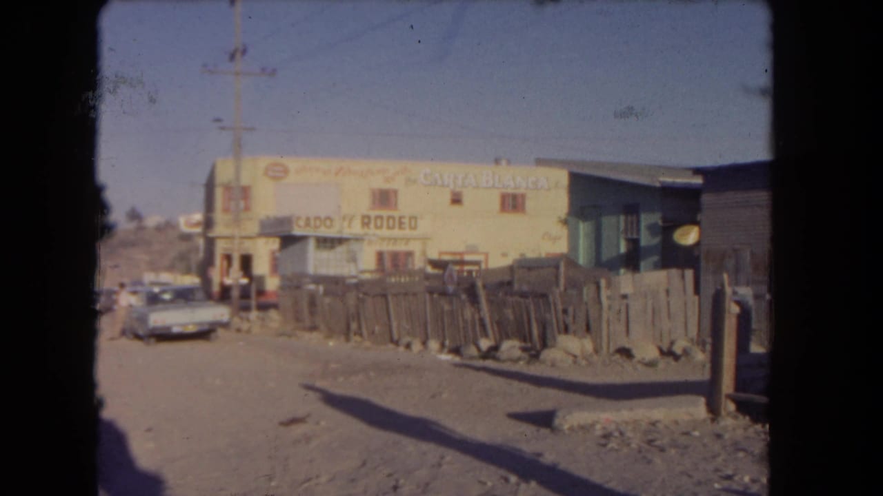 1962 Los Angeles 8mm frame of the same dusty roadside after the man has moved out of view, with a yellow building, rough fence, utility pole, and parked car visible.