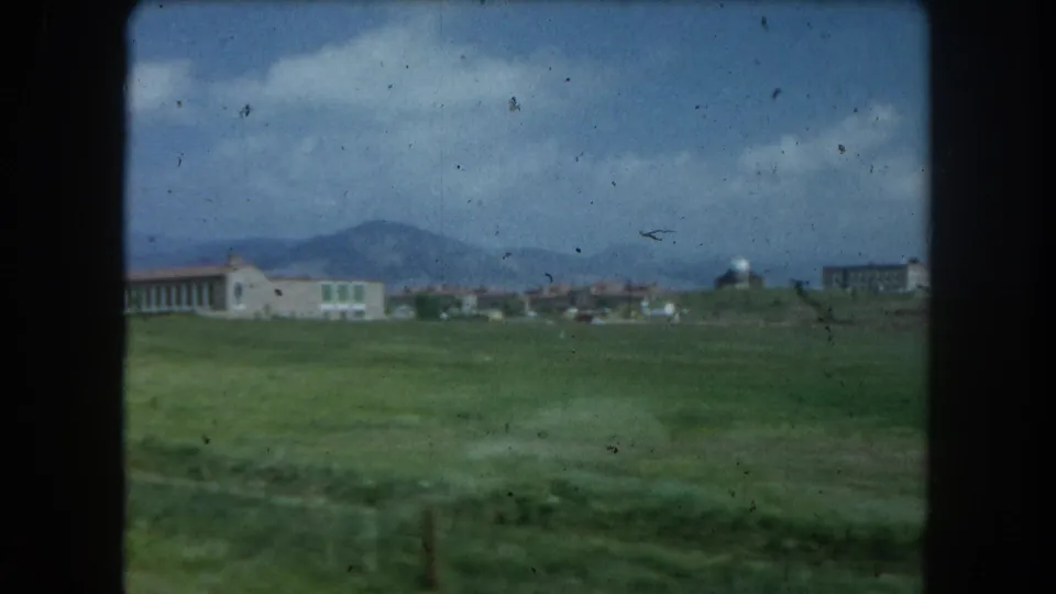 1962 Summit County, Colorado view of a green valley with low buildings, cloudy sky, and mountains in the distance.