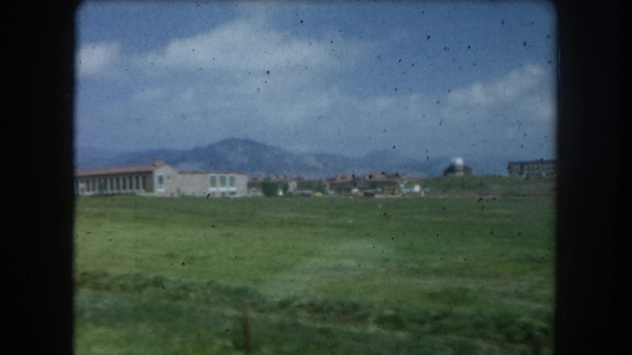 8mm home-movie frame circa 1962 showing a green field and low institutional building near Summit County, Colorado, with mountain foothills under a partly cloudy sky.