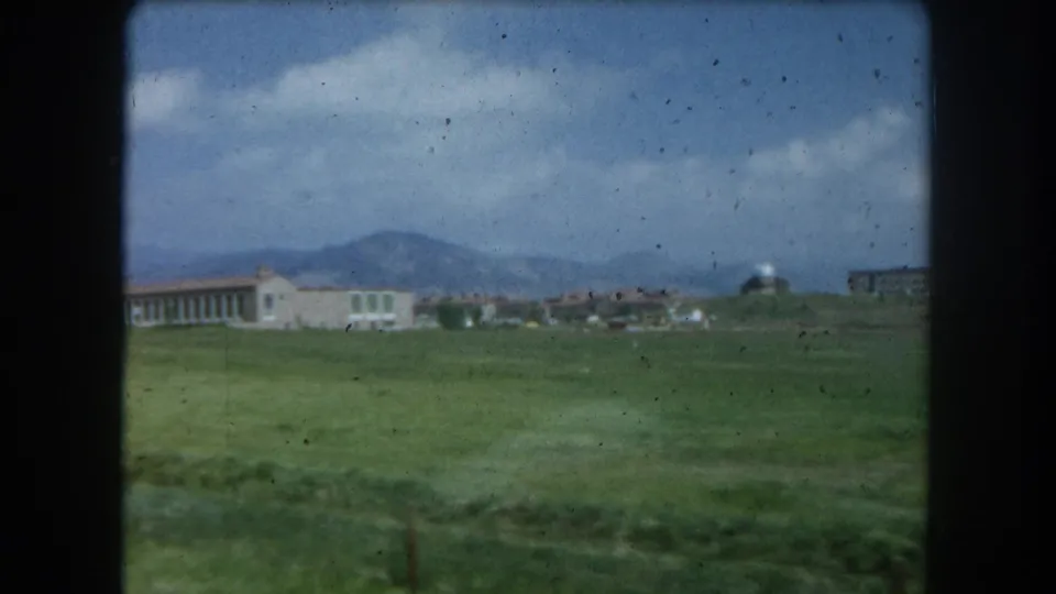 8mm home-movie frame circa 1962 showing a green field and low institutional building near Summit County, Colorado, with mountain foothills under a partly cloudy sky.