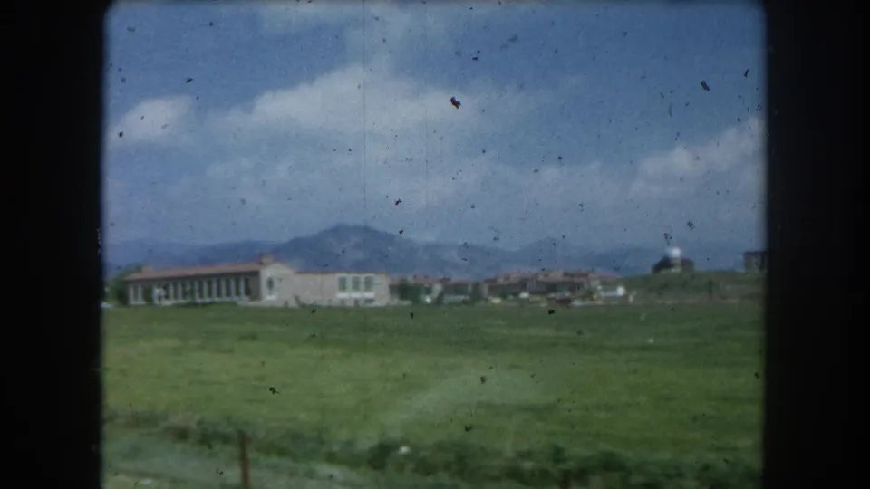 8mm color home-movie frame circa 1962 continuing the driving-past view of a meadow and building near Summit County, Colorado, with mountain backdrop.