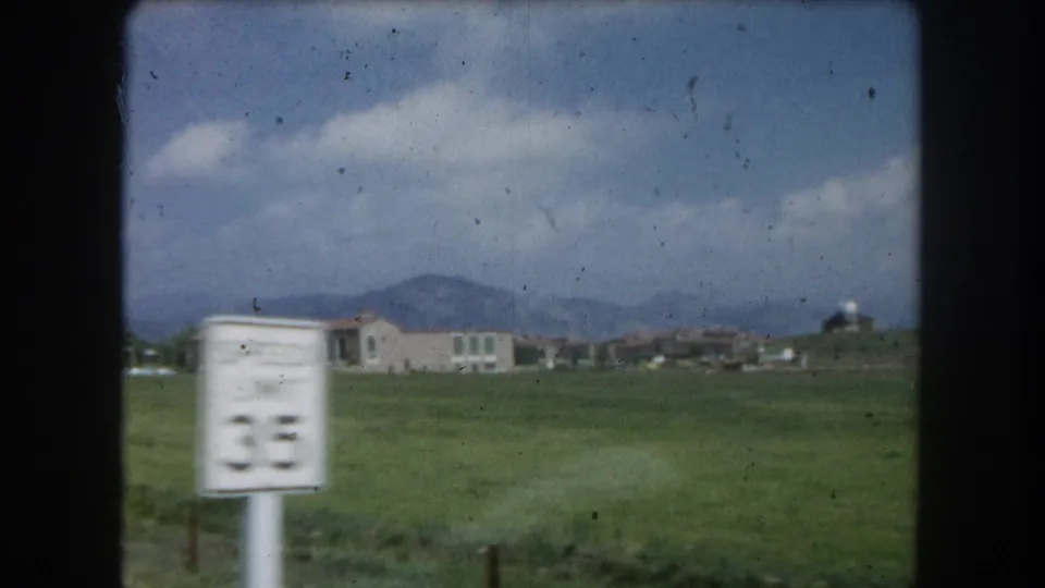 8mm home-movie frame circa 1962: a 35 mph speed limit sign on a Colorado roadside with a low building complex and mountain foothills behind a green field.