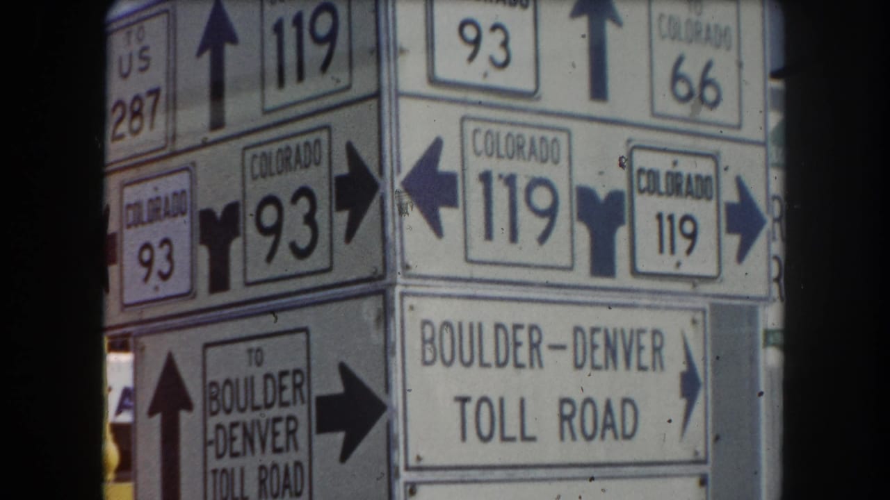 Roadside signpost cluster with Colorado routes 93, 119, 66, US 287, and Boulder-Denver Toll Road signs; red vehicle partially visible at frame left, circa 1962 Summit County, Colorado.