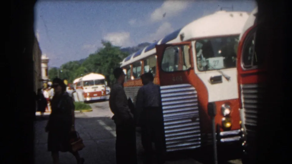 Bus stop scene in a Colorado mountain town circa 1962: red and white inter-city bus in the foreground, a second bus behind, pedestrians including one figure in a hat, tree-lined street, and mountain terrain under blue sky.