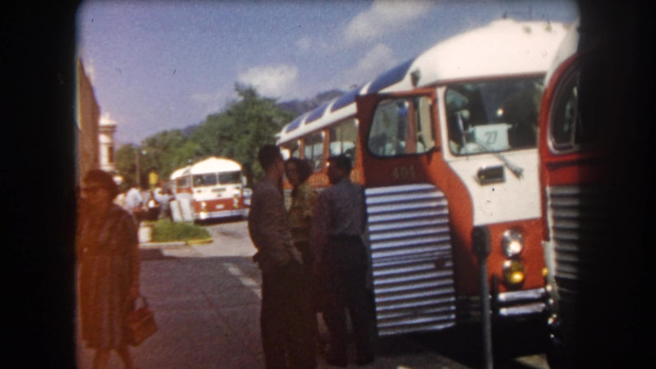 Red and white intercity coaches parked on a street in what appears to be Summit County, Colorado, circa 1962; passengers in early-1960s dress boarding or waiting; mountains and trees behind.