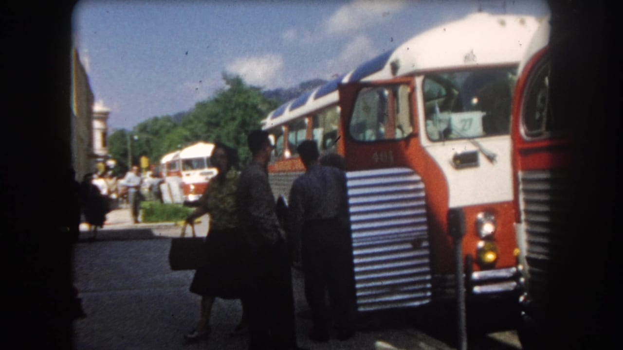 Woman with handbag pauses near the front of a red-and-white intercity coach in what appears to be Summit County, Colorado, circa 1962; mountain backdrop and summer trees visible.