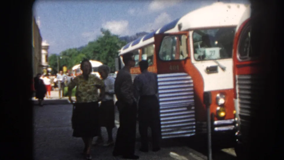 Group of passengers in early-1960s clothing stand on a street in what may be Summit County, Colorado, circa 1962; red-and-white coach, vehicles, and mountain backdrop visible.