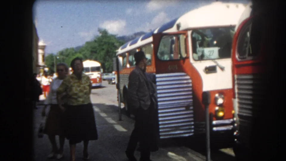 Two people in 1960s dress stand near the front of a red-and-white intercity coach, likely Summit County, Colorado, circa 1962; mountain backdrop and second bus visible.