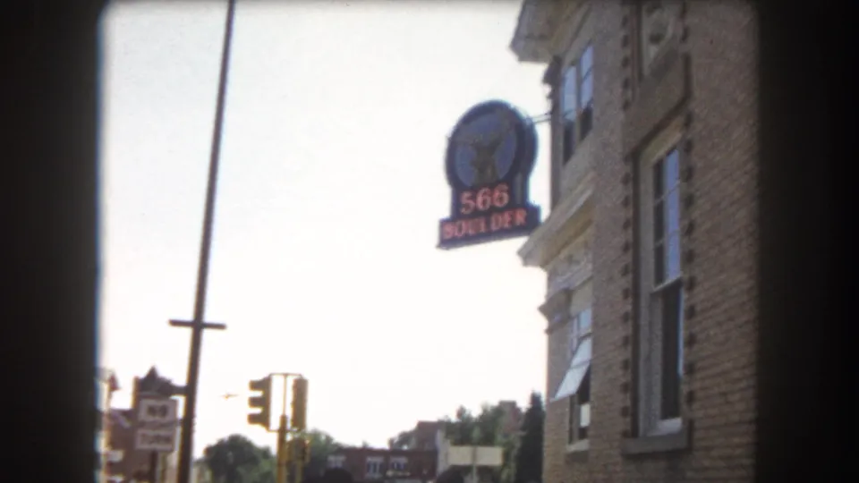 8mm home movie frame showing a brick commercial corner building with a prominent '566 BOULDER' sign and traffic light, possibly Boulder, Colorado, circa 1962.