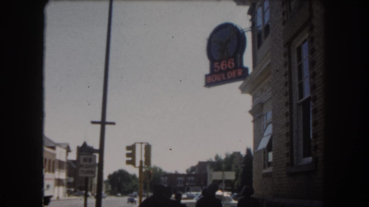8mm street view of a downtown Colorado intersection showing the '566 BOULDER' brick corner building, traffic signal, utility poles, and early-1960s automobiles, circa 1962.