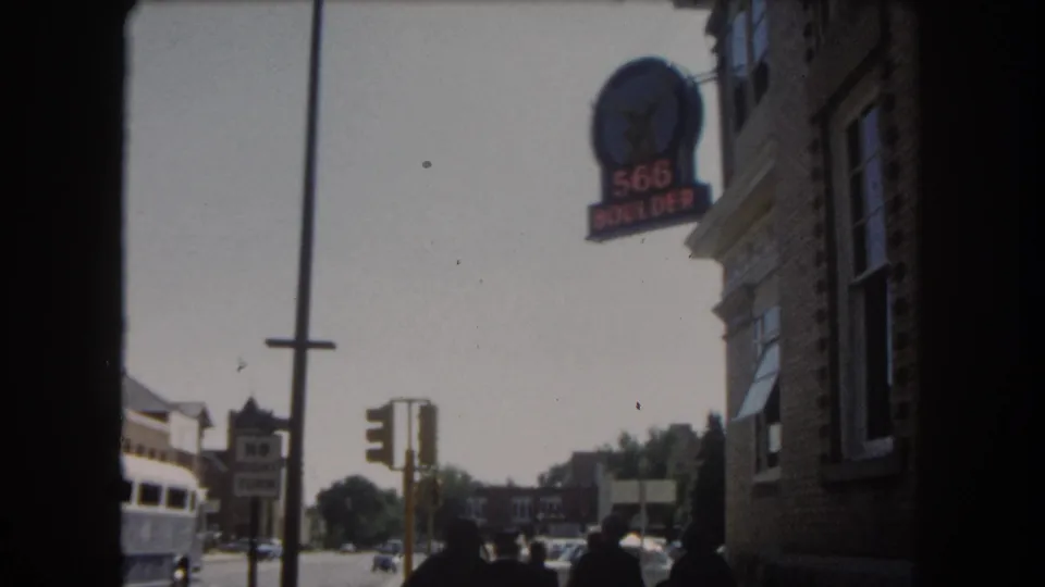 8mm frame showing the brick corner building with '566 BOULDER' sign, a transit bus at lower left, and pedestrians on a sunny Colorado street, circa 1962.