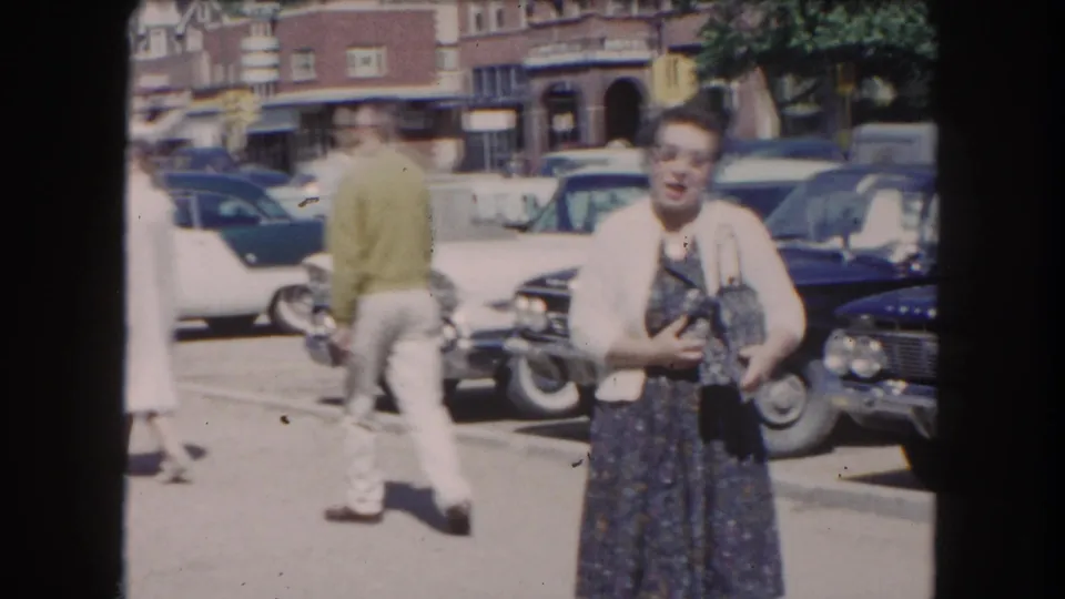 8mm close-up of a woman in cat-eye glasses, cardigan, and patterned dress walking toward camera on a sunny Colorado street lined with early-1960s American automobiles, circa 1962.