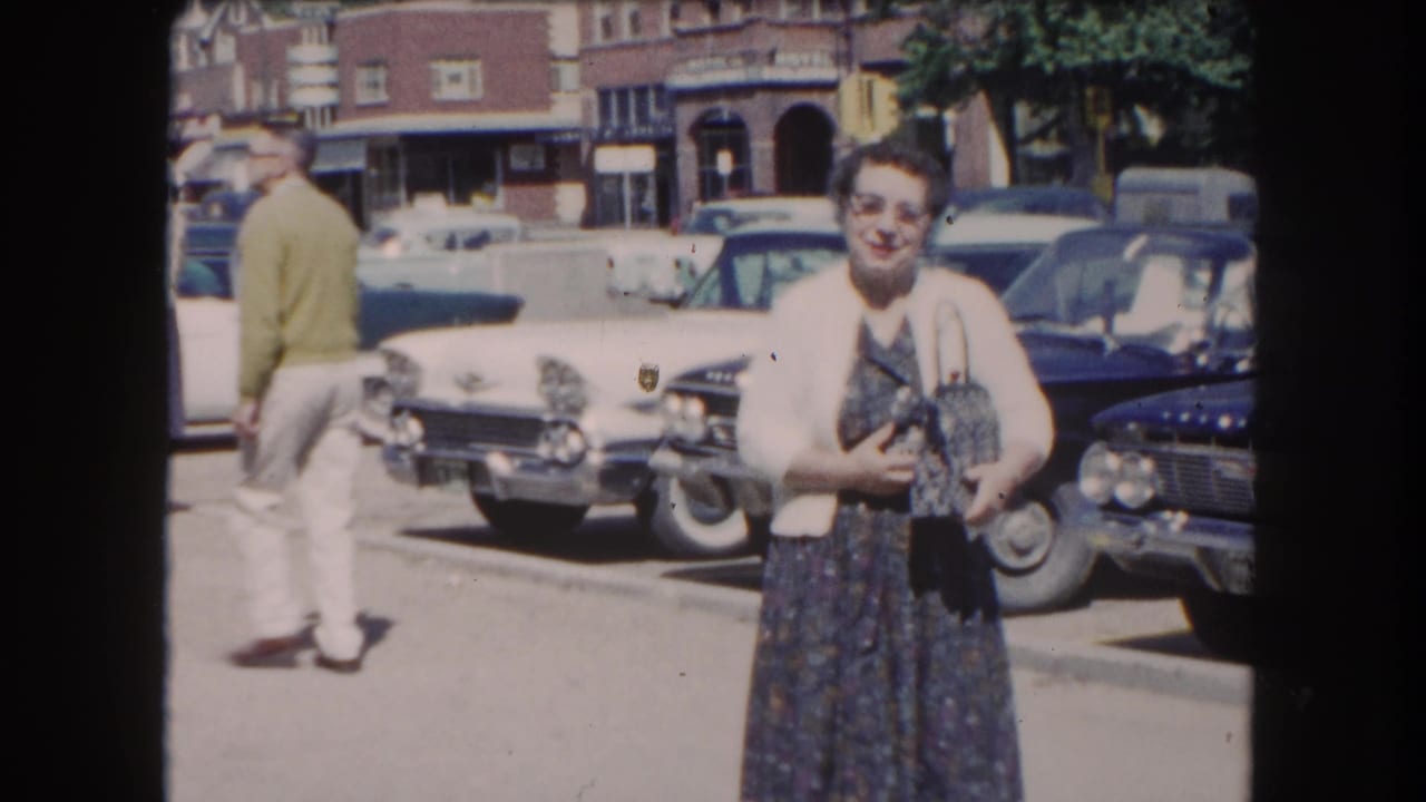 A woman in early-1960s dress stands in a sunlit parking lot, possibly in a small Colorado town circa 1962; classic American automobiles line the background.