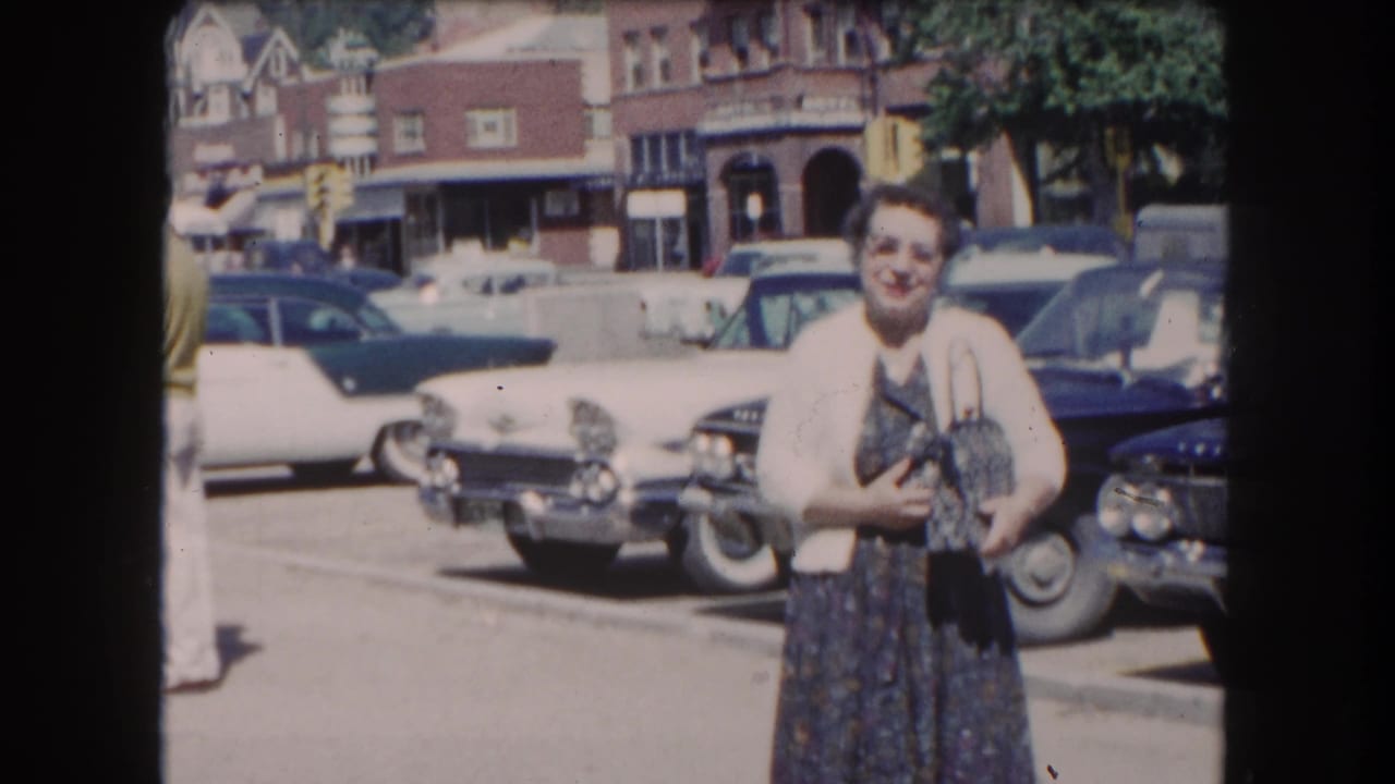 Woman in a white cardigan and floral dress poses in a parking lot circa early 1960s; a white late-1950s/early-1960s American automobile with prominent tail fins is visible behind her.