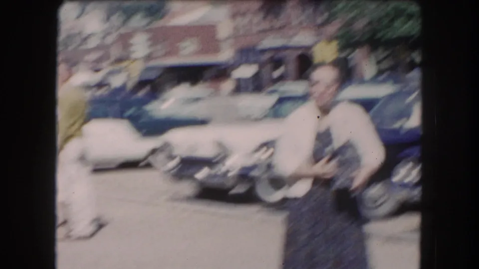 Motion-blurred 8mm frame showing a woman in a white cardigan and a man in a green jacket crossing a parking lot, circa early 1960s; camera movement renders most details indistinct.