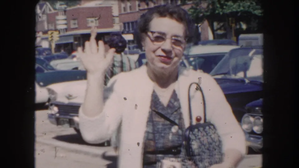 Close-up of an older woman in cat-eye glasses and a white cardigan waving at the camera in a parking lot lined with early-1960s American automobiles, circa 1962.
