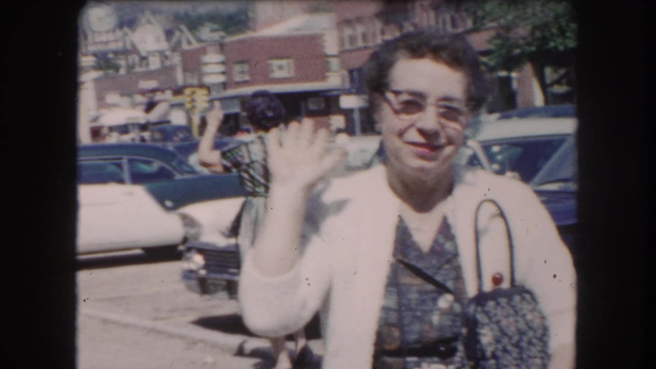 Older woman in glasses and cardigan waves and smiles at the camera; early-1960s American cars and a small-town brick streetscape are visible in the background, Summit County Colorado area, c.1962.