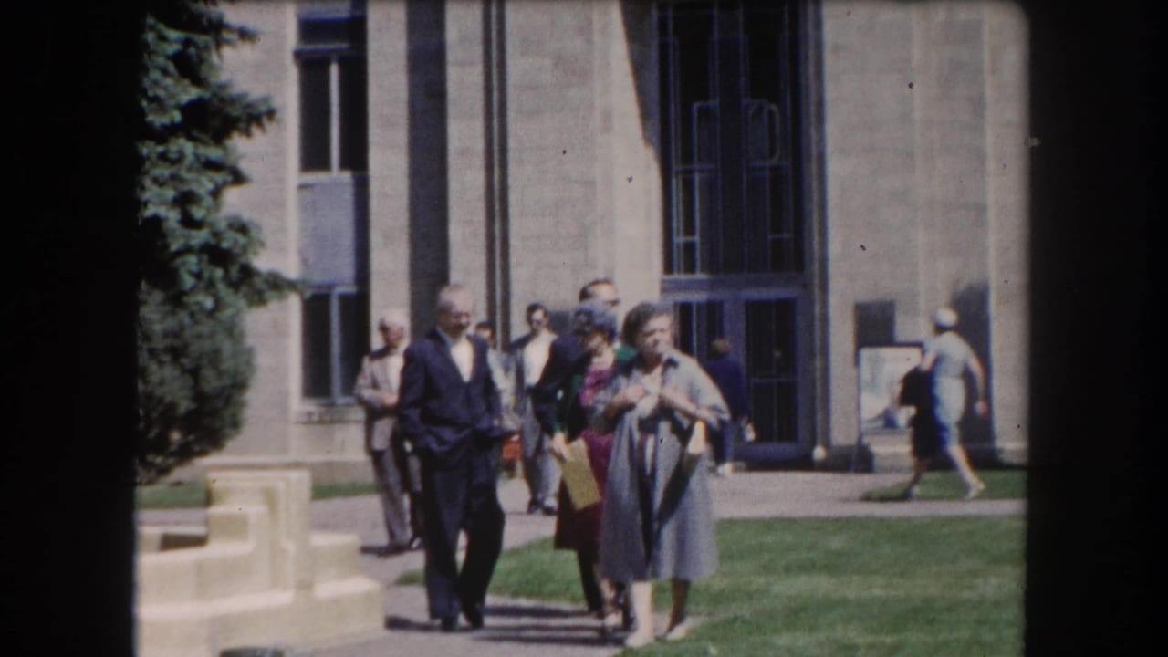 Small group of adults, men in suits and women in dresses, walking toward the camera on a sunny path beside a stone civic building, Summit County Colorado area, c.1962.