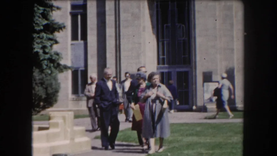 Small group of adults, men in suits and women in dresses, walking toward the camera on a sunny path beside a stone civic building, Summit County Colorado area, c.1962.