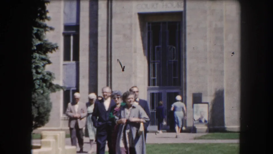 Several adults in 1960s clothing walk before the stone entrance of a courthouse with carved lettering; evergreen tree at left, Summit County Colorado area, c.1962.