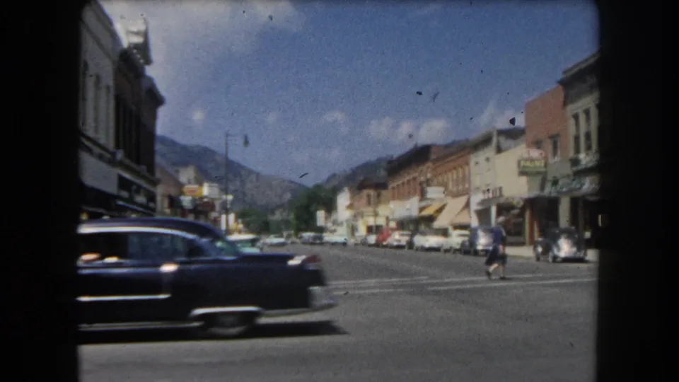 Dark early-1960s American sedan passing storefronts on what appears to be the main street of a Colorado mountain town, circa 1962.