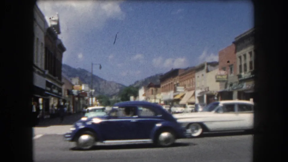Dark blue early-1960s sedan and white station wagon on the main commercial street of a Colorado mountain town, circa 1962, with peaks visible behind the buildings.