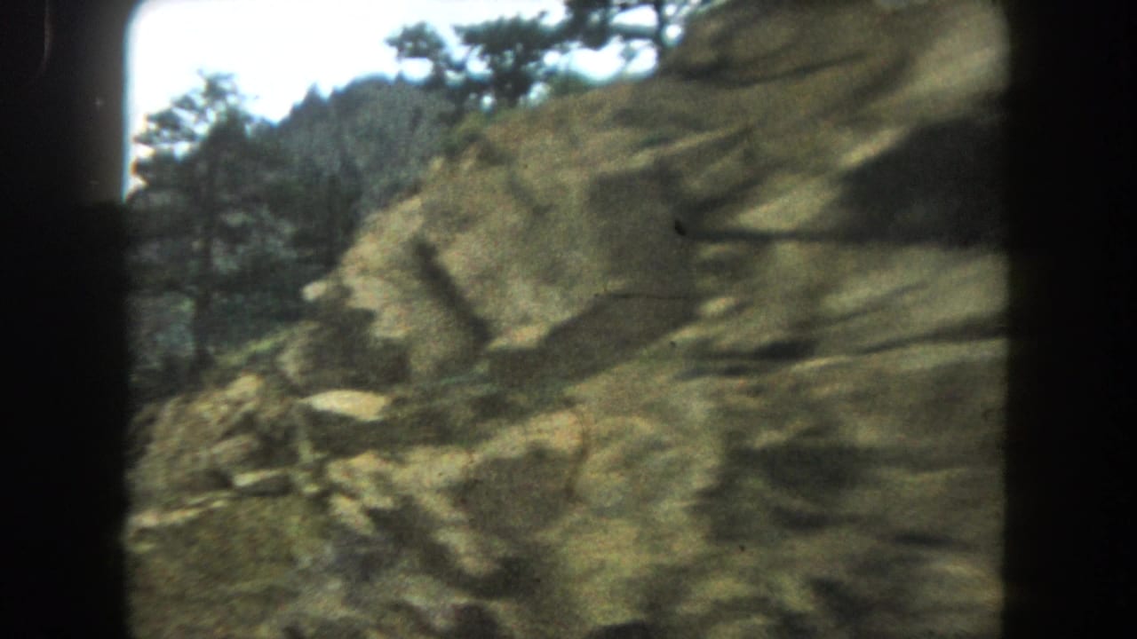 8mm home movie frame of a rocky mountain face with a pine tree at upper right, likely Colorado high country, circa 1962.