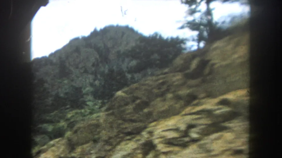 8mm home movie frame showing a mountain ridgeline above a dense pine forest, filmed from a moving car in Colorado, circa 1962.