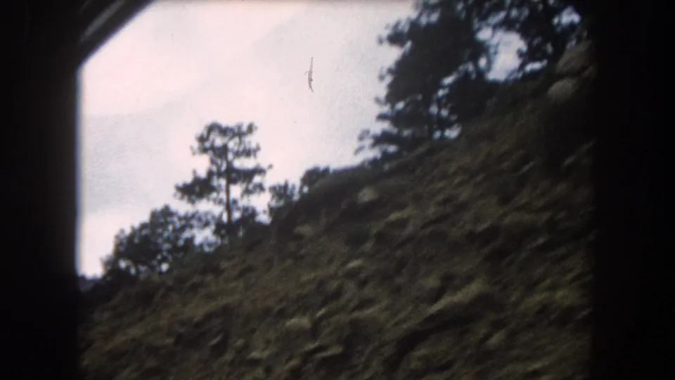 8mm home movie frame circa 1962, steep rocky slope with sparse pine trees against pale sky, possibly Summit County, Colorado
