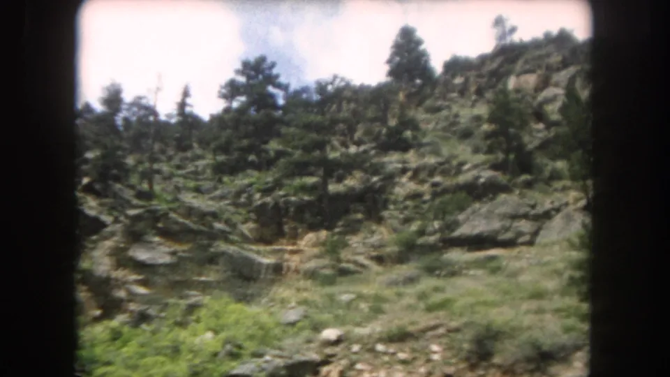 Close view of a rocky Colorado mountain slope with sparse pine trees and exposed rock face, captured from a moving train on 8mm film, circa 1962.