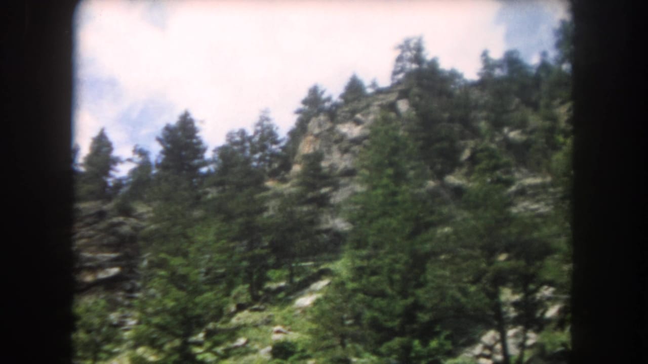 Rocky mountain slope densely forested with conifers under a partly cloudy sky, 8mm film.