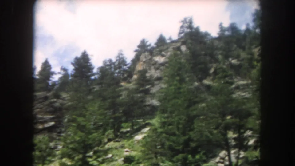8mm film frame of a rocky Colorado mountain slope covered in conifer trees with granite outcrops and a partly cloudy sky, circa 1962.