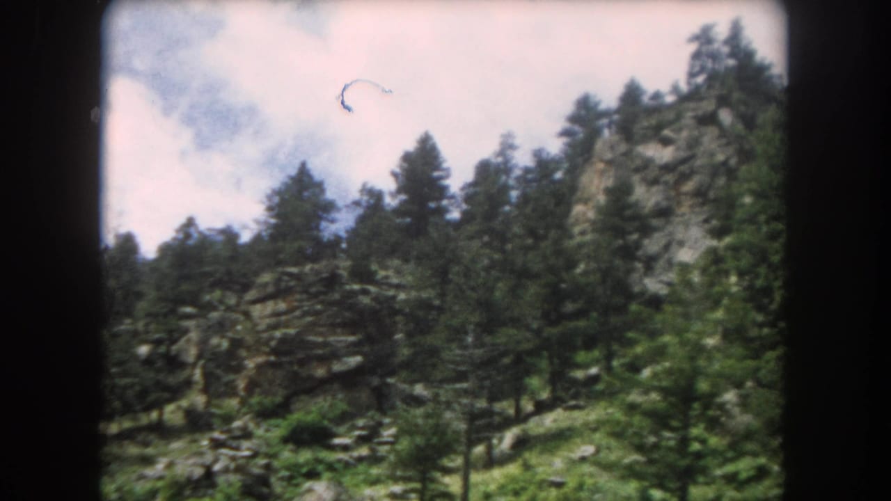 Exposed granite rock face flanked by conifers under partly cloudy sky, with a small circular film artifact visible near upper centre, 8mm film.