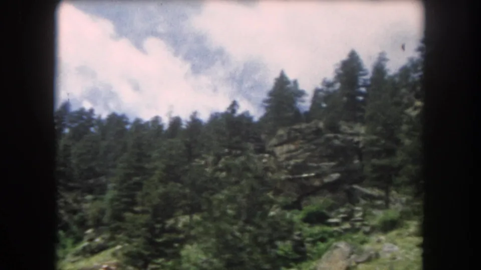 8mm film frame of a conifer treeline against a partly cloudy mountain sky with a rocky forested slope in the foreground, Colorado Rockies, circa 1962.