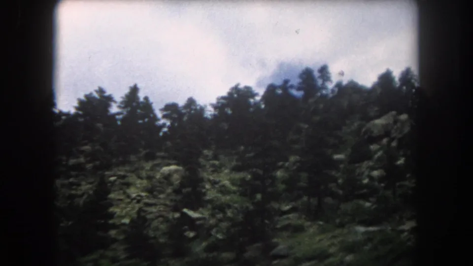 8mm film frame of a dense conifer forest silhouetted against a bright partly cloudy sky in the Colorado Rockies, with rocky terrain visible below, circa 1962.