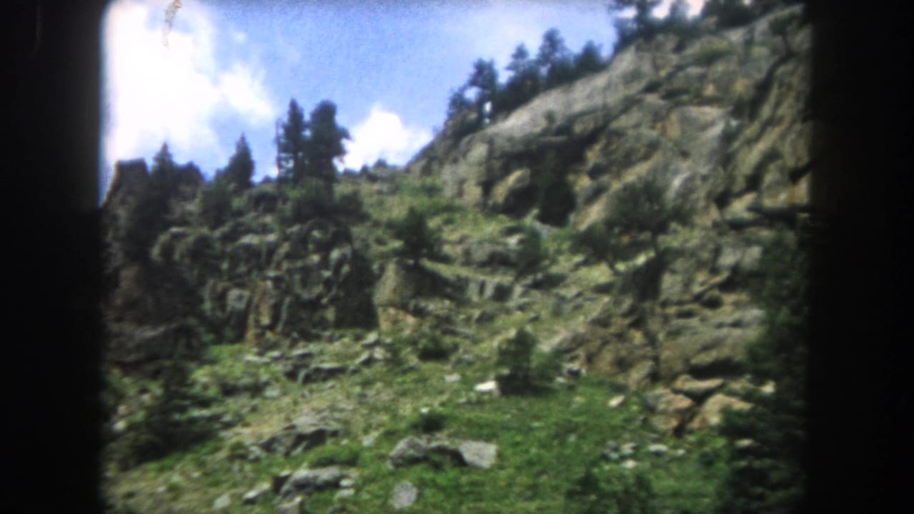 8mm home movie frame from 1962 showing a rocky mountain slope with granite outcroppings, conifers, and patchy grass, Summit County, Colorado.
