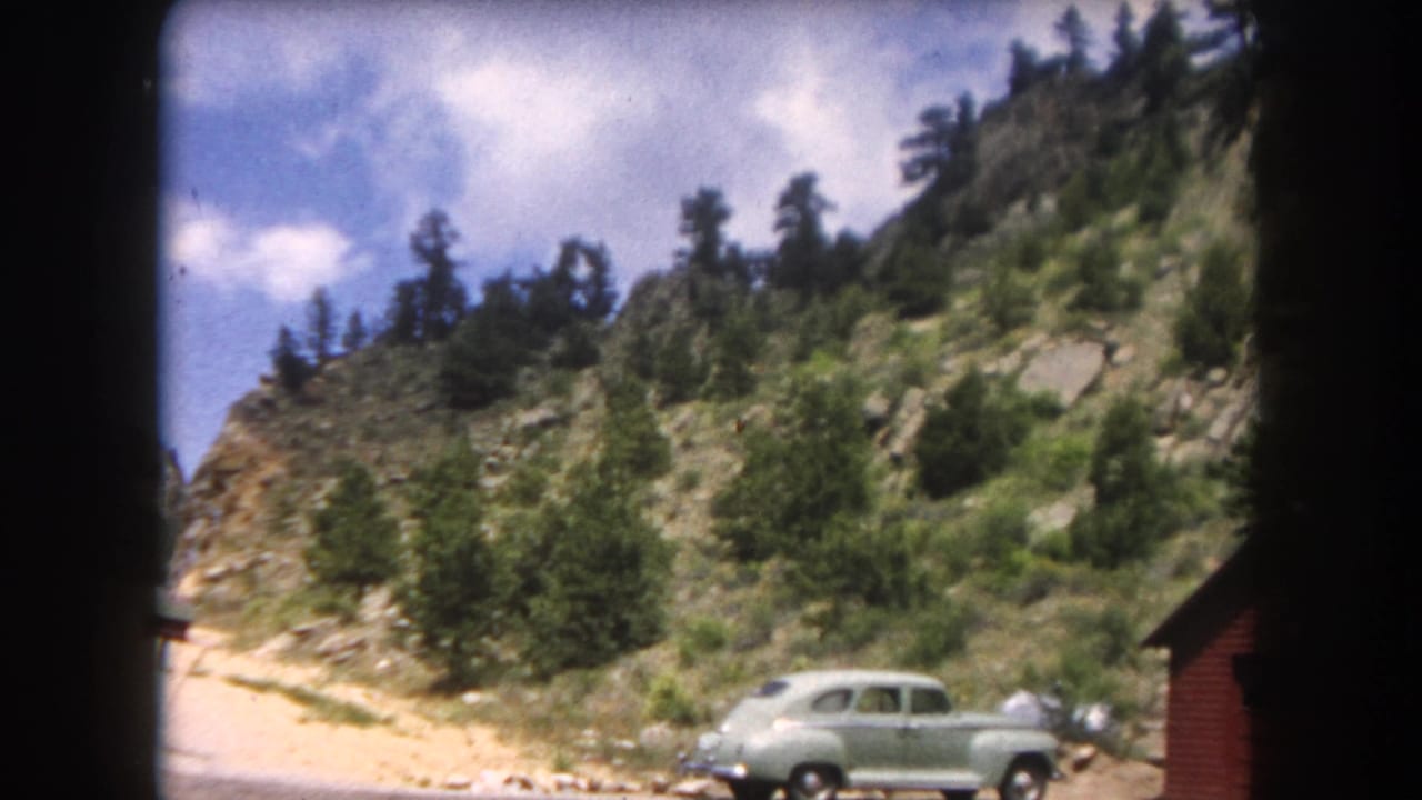 A rounded light-green mid-century sedan on a gravel pull-off in what appears to be Summit County, Colorado, circa 1962, with a red cabin and forested rocky slope.