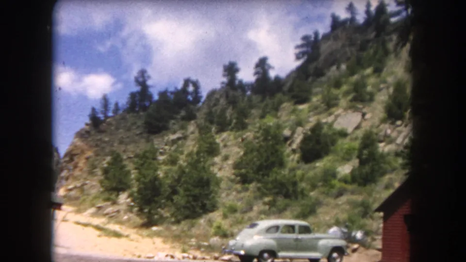 A rounded light-green mid-century sedan on a gravel pull-off in what appears to be Summit County, Colorado, circa 1962, with a red cabin and forested rocky slope.