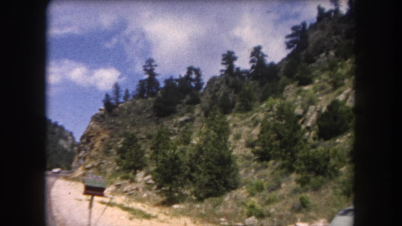 A mountain road through rocky terrain dense with conifers, likely Summit County, Colorado, circa 1962; red cabin visible at the road's edge.