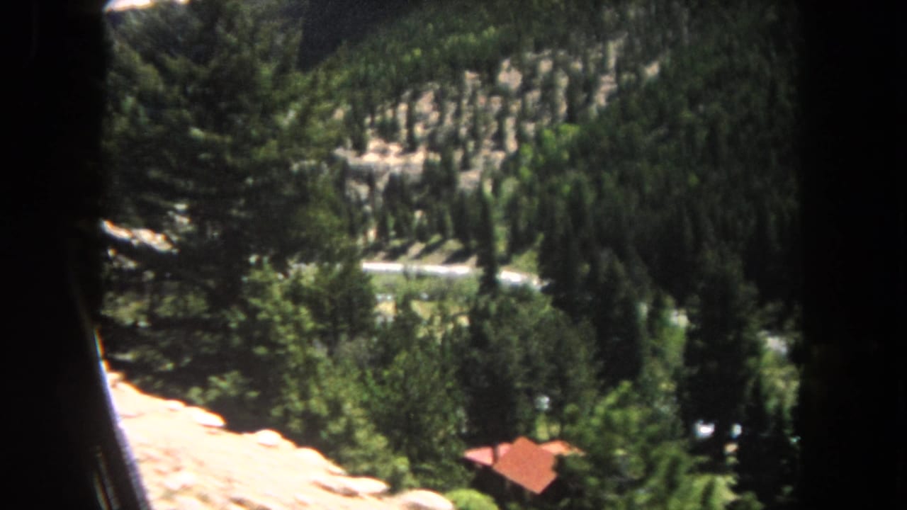 8mm home movie frame circa 1962, looking down a pine-forested slope in Summit County, Colorado, with a small red-roofed cabin in the middle distance.