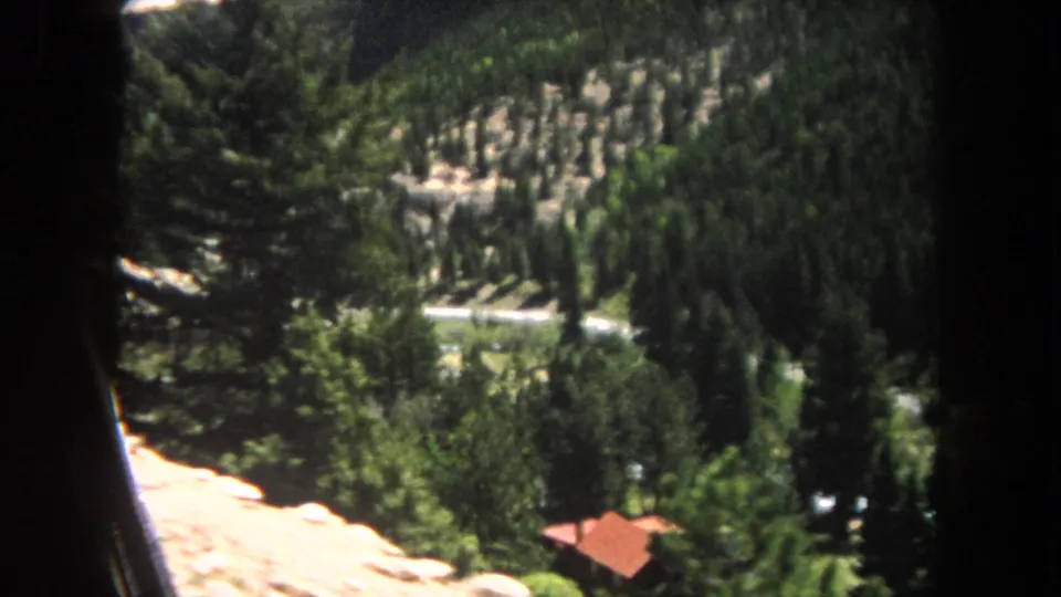 8mm home movie frame circa 1962, looking down a pine-forested slope in Summit County, Colorado, with a small red-roofed cabin in the middle distance.