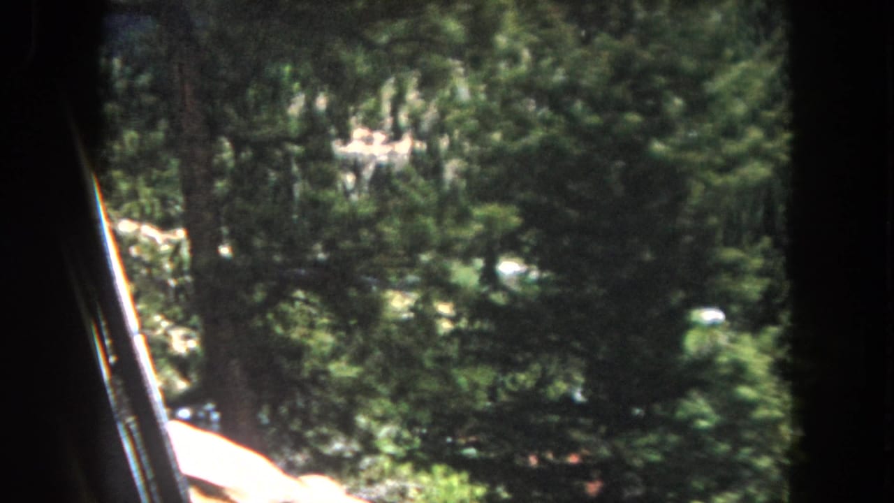 8mm home movie frame circa 1962, Summit County, Colorado: pine forest covering a steep valley slope with a red-roofed building visible in the middle distance at right frame edge.