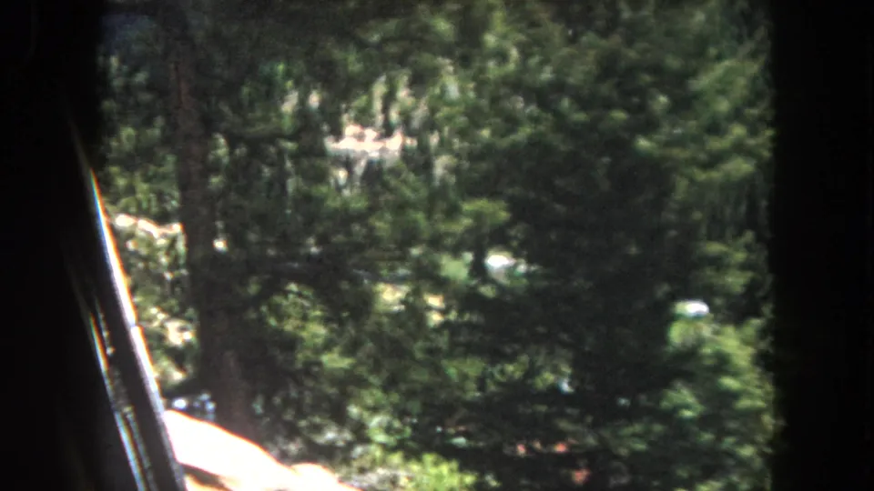 8mm home movie frame circa 1962, Summit County, Colorado: pine forest covering a steep valley slope with a red-roofed building visible in the middle distance at right frame edge.