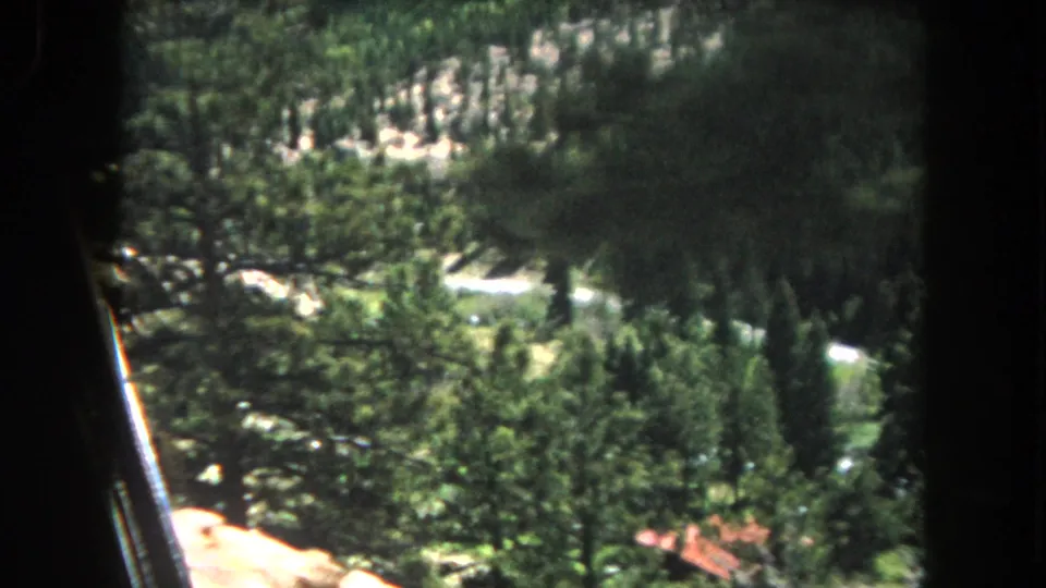 8mm home movie frame circa 1962, Summit County, Colorado: mountain drive POV showing rocky outcrops below the tree canopy and a valley visible in the lower portion of the frame.