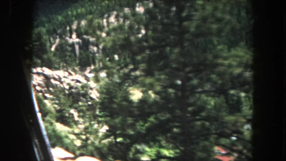 8mm home movie frame circa 1962, Summit County, Colorado: closing frames of a mountain drive showing dense conifers, exposed rock faces, and a deep valley through the film-gate vignette.