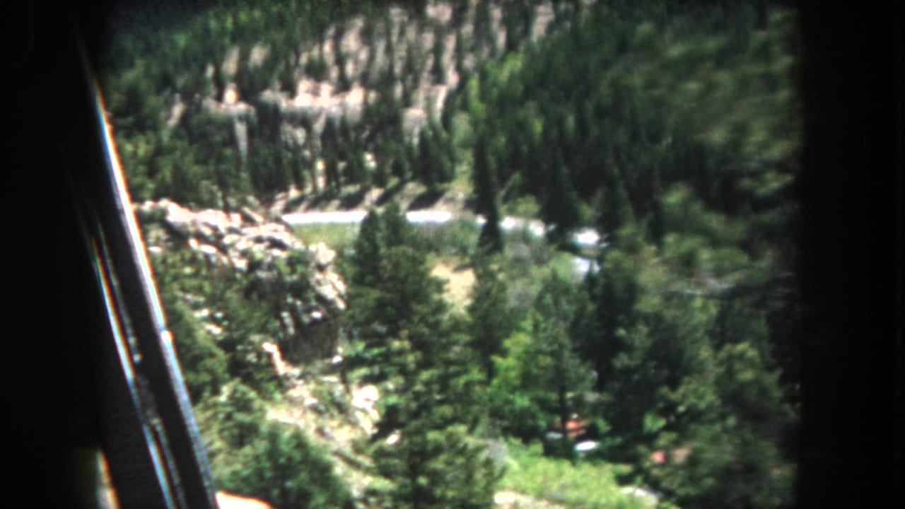 View from inside a moving vehicle looking steeply down a forested mountain valley with evergreen trees, rocky hillside, and a valley floor visible below.