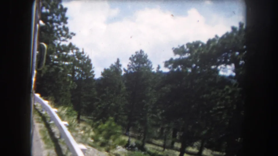8mm home movie circa 1962 of a mountain road with white guardrail, pine trees, and cumulus clouds, likely in the Colorado Rocky Mountains.