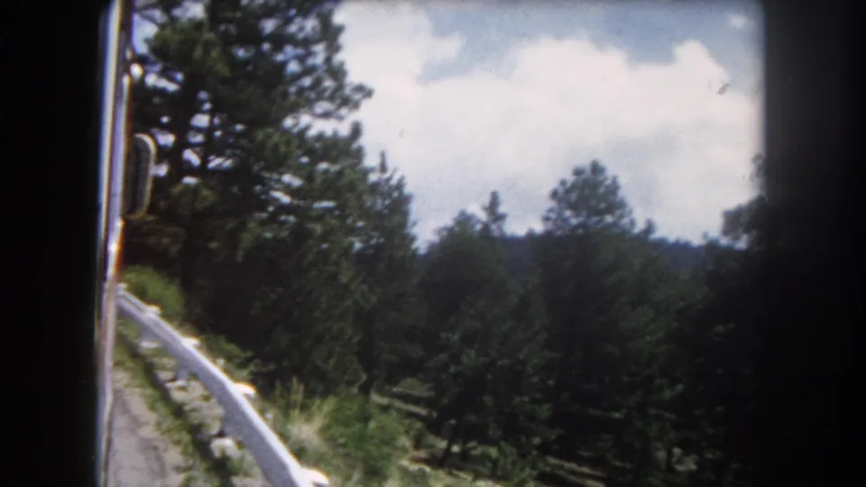 8mm home movie circa 1962 of a mountain road lined with pine trees and a white guardrail under a partly cloudy sky, consistent with the Colorado Rocky Mountains.