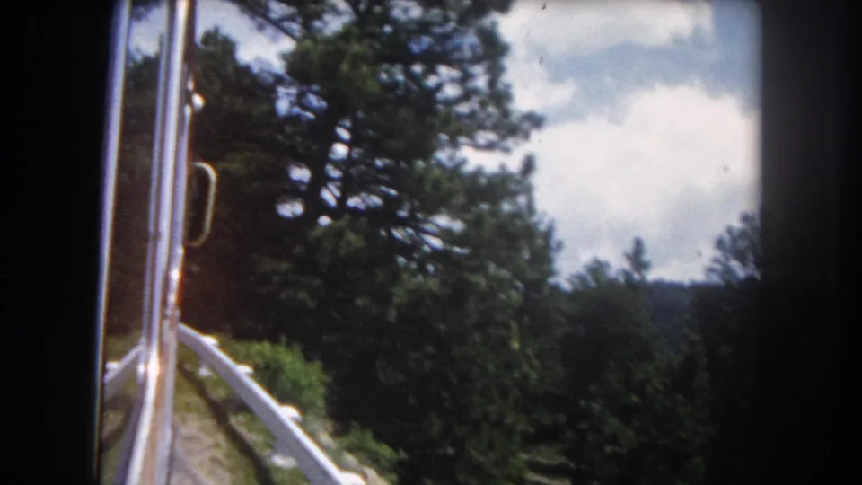 8mm color shot from a moving vehicle window showing dense pine forest and white metal guardrail on a Summit County, Colorado mountain road, circa 1962.