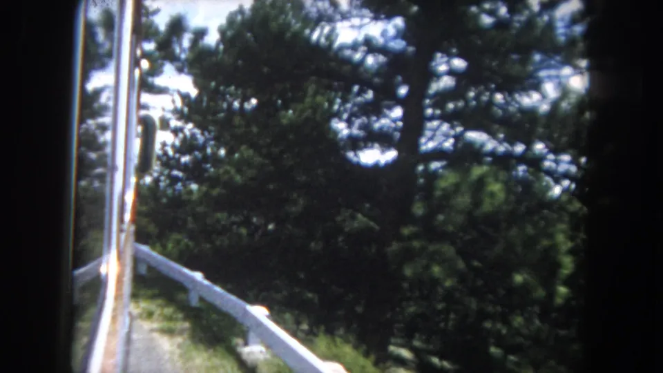 8mm color footage from a moving vehicle showing pine forest and white mountain-road guardrail under a partly cloudy sky, consistent with early 1960s Colorado Rocky Mountain road travel.